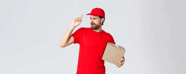 Entrega de pedidos, compras online e conceito de envio de pacotes. Carefree mensageiro barbudo bonito em uniforme vermelho, boné e t-shirt, segurando pacote caixa, canto canção, fundo cinza — Fotografia de Stock