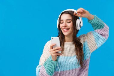 Mutlu karizmatik ve kaygısız güler yüzlü Avrupalı kadın yeni yılbaşı hediyesi olarak alınan yeni kulaklıklarda müzik kalitesinin tadını çıkarıyor, akıllı telefon ve mavi arka plan ile el kaldırıyor.