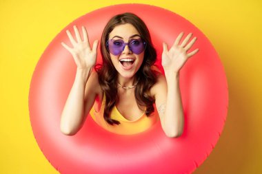 Close up portrait of enthusiastic young woman inside pink swimming ring, laughing and smiling, enjoying beach holiday, summer vacation, yellow background