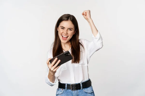 Happy brunette woman playing mobile video game, smiling and looking at screen excited, standing over white background — Stockfoto