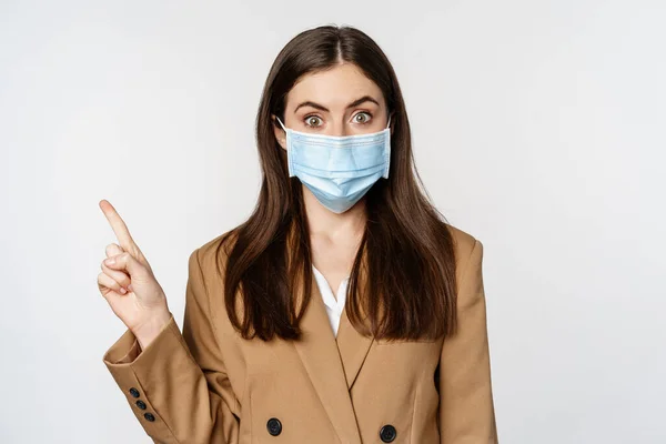 Coronavirus and people concept. Portrait of business woman at workplace wearing face mask, pointing finger left at logo, company banner, white background — стоковое фото