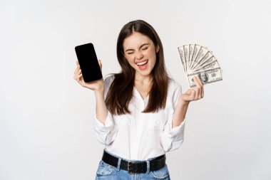 Enthusiastic young woman winning money, showing smartphone app interface and cash, microcredit, prize concept, standing over white background