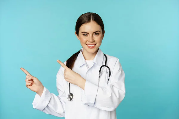 Portrait of smiling medical worker, girl doctor in white coat with stethoscope, pointing fingers left, showing medical clinic advertisement, torquoise background — Fotografia de Stock