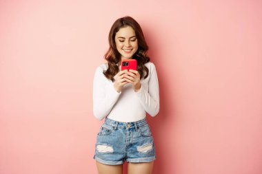 Smiling brunette girl using smartphone, texting message, order online on mobile phone app, standing over pink background
