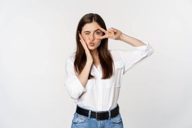 Beautiful girl with healthy skin, showing peace sign on face and winking, standing over white background. People and emotions concept
