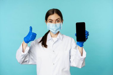 Portrait of doctor in medical face mask and gloves, showing mobile phone app, smartphone screen and thumb up, recommending online checkup website, standing over blue background