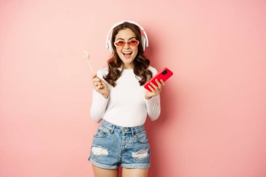 Stylish summer girl dancing with smartphone and lolipop, listening music in headphones, standing in sunglasses against pink background