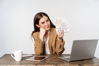 Portrait of businesswoman sitting in office with money, working and making profit income, posing happy against white studio background