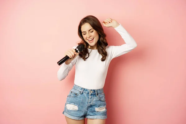 Karaoke. Happy smiling girl singing in microphone, dancing and having fun, standing over pink background — ストック写真