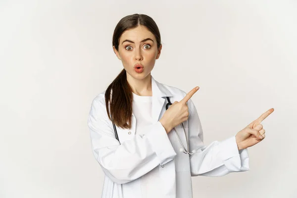 Woman doctor, physician in medical uniform, pointing fingers right, showing banner advertisement, smiling amazed, checking out promo discount, white background — Fotografia de Stock