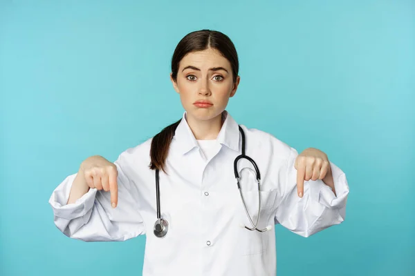 Sad and gloomy doctor, female medical worker pointing fingers down, looking upset, showing bad news, standing in white coat over blue background — ストック写真