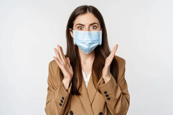 Workaplce and pandemic concept. Shocked business woman in face medical mask, gasping, looking startled and concerned at camera, standing over white background — Fotografia de Stock