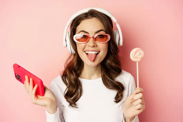 Beautiful female model listening music in headphones, holding lolipop and mobile phone, posing in sunglasses, standing over pink background — Zdjęcie stockowe