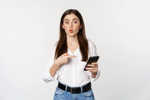 Young woman pointing at mobile phone while looking interested in app, showing smth on smartphone, standing over white background — стоковое фото