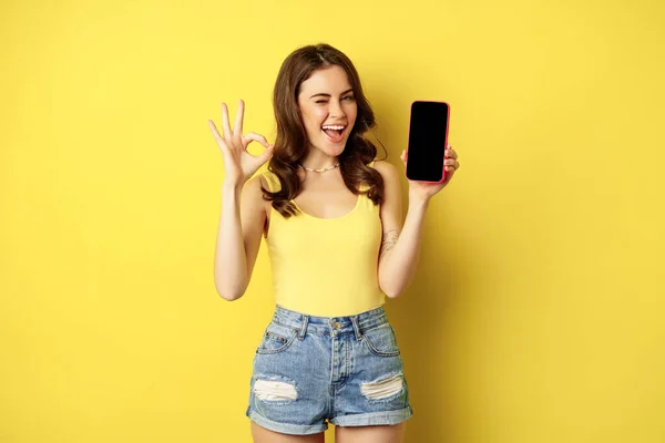 Pretty brunetka kobieta, pokazując ekran telefonu komórkowego, smartphone interfejs aplikacji i dobrze znak, polecam stronę internetową, sklep, stojąc na żółtym tle — Zdjęcie stockowe