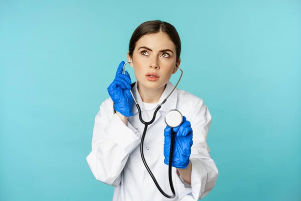 Serious looking woman doctor listening in stethoscope with focused face, checkup for client, standing over torquoise background — Zdjęcie stockowe