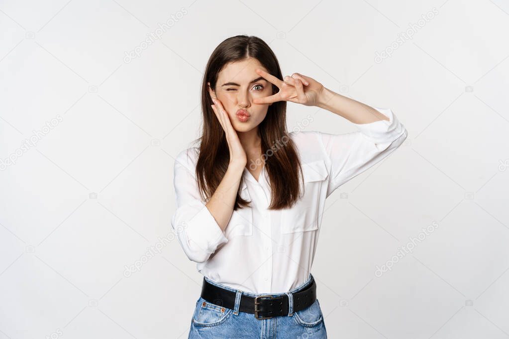 Beautiful girl with healthy skin, showing peace sign on face and winking, standing over white background. People and emotions concept.