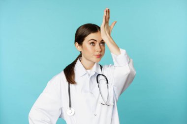 Portrait of annoyed, tired woman doctor, facepalm, roll eyes frustrated, bothered by smth stupid, standing in white coat over torquoise background