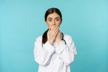 Shocked and concerned woman doctor, clinic worker, covering mouth with hands, looking startled at camera, standing over blue background