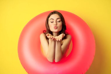 Portrait of beautiful female model posing with pink swimming beach ring, sending air kiss, mwah flirty, standing over yellow background