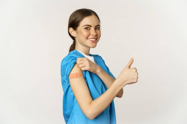 Smiling nurse, doctor in scrubs, medical worker showing her vaccinated shoulder with patch, thumbs up, recommending vaccinate from coronavirus, white background