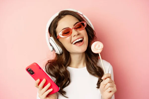 Beautiful female model listening music in headphones, holding lolipop and mobile phone, posing in sunglasses, standing over pink background — Zdjęcie stockowe