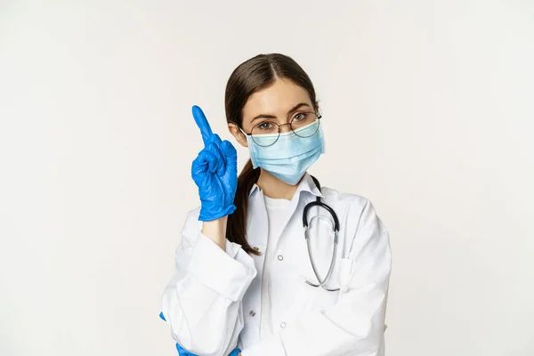 Närbild porträtt av kvinnlig läkare i glasögon, medicinsk ansiktsmask och handskar, peka finger upp på länk, klinik logotyp, reklam på toppen, står över vit bakgrund — Stockfoto