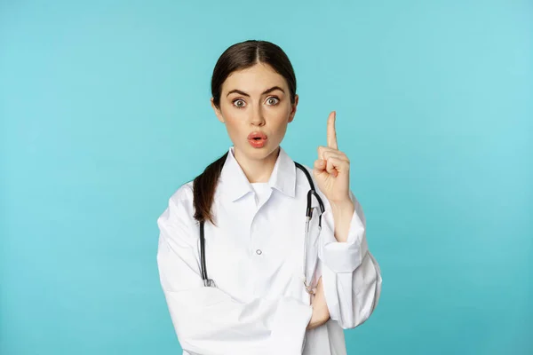 Young doctor, woman physician in white coat raising finger, pointing up, suggesting smth, has solution, revelation, standing over torquoise background — Zdjęcie stockowe