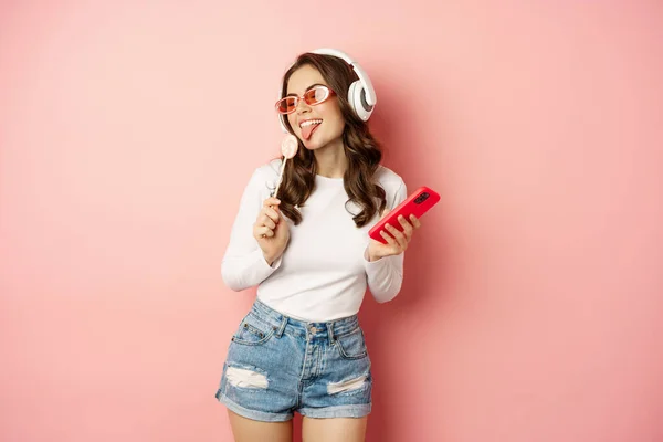 Elegante chica de verano bailando con smartphone y lolipop, escuchando música en auriculares, de pie en gafas de sol sobre fondo rosa — Foto de Stock