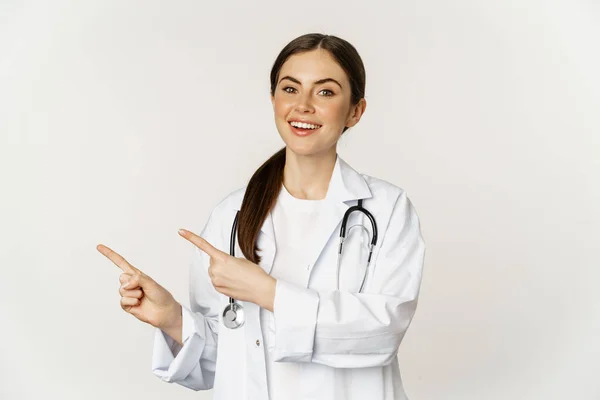Portrait of smiling young woman doctor, healthcare medical worker, pointing fingers left, showing clinic promo, logo or banner, standing over white background — Zdjęcie stockowe