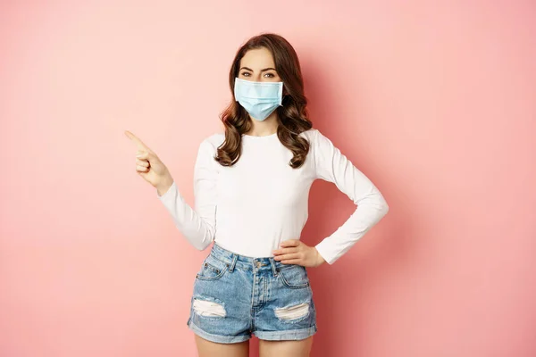 Beautiful stylish woman in medical face mask from covid, pointing finger at upper left corner, showing advertisement, standing over pink background — Φωτογραφία Αρχείου