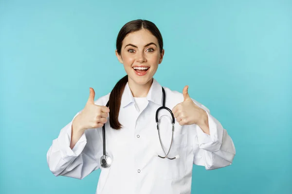 Boldog nő orvos, egészségügyi dolgozó fehér köpenyben, mutatja hüvelykujját fel a jóváhagyást, mint valami, dicséret, áll a kék háttér felett — Stock Fotó