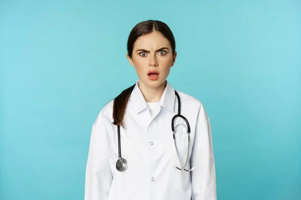 Retrato de doctora conmocionada, pasante del hospital con bata blanca, mirando preocupada y confundida a la cámara, cara de incredulidad, de pie sobre fondo turquesa — Foto de Stock