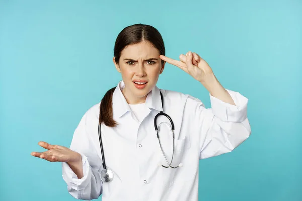 Médico mujer molesta señalando con el dedo a la cabeza, regañando a alguien estúpido, loco o extraño, de pie sobre fondo de color turquesa — Foto de Stock