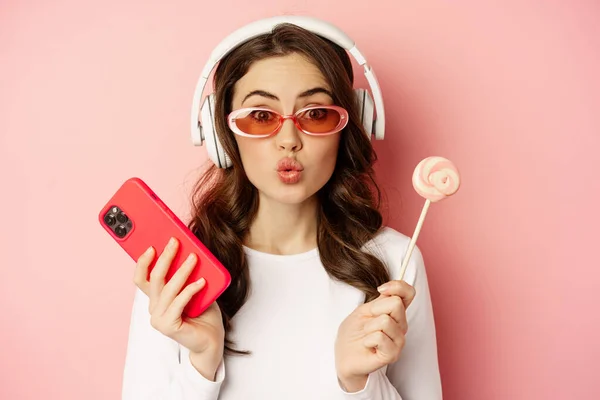 Stylish young woman with lolipop and cellphone, wearing sunglasses and headphones, listening music, standing over pink background — Zdjęcie stockowe
