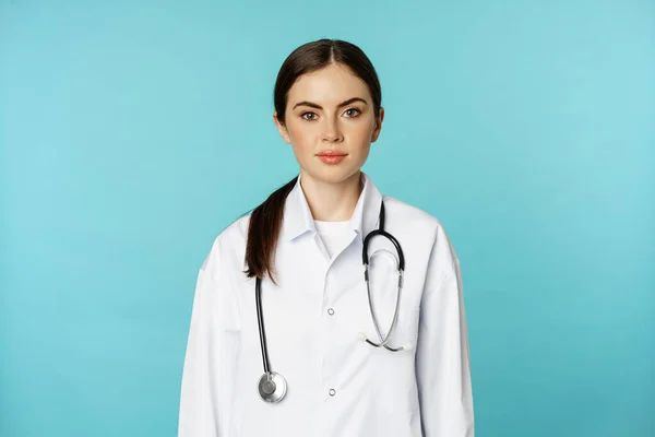 Medical staff and doctors concept. Young smiling female doctor, healthcare worker in white coat and stethoscope, looking confident, waiting patients, blue background — Photo