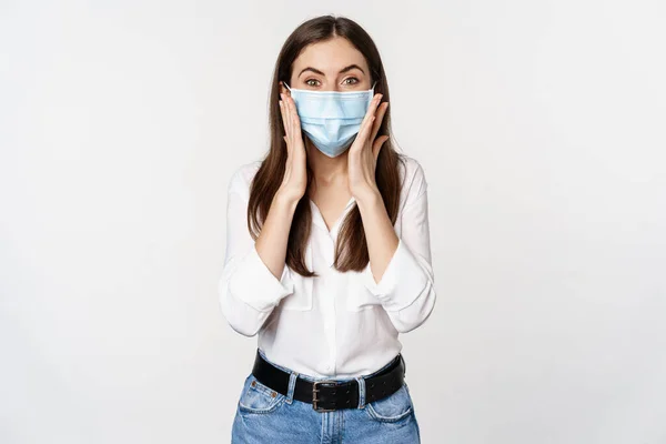 Surprised woman in face medical mask looking amazed, checking out awesome news, big reveal, standing over white background — Foto de Stock