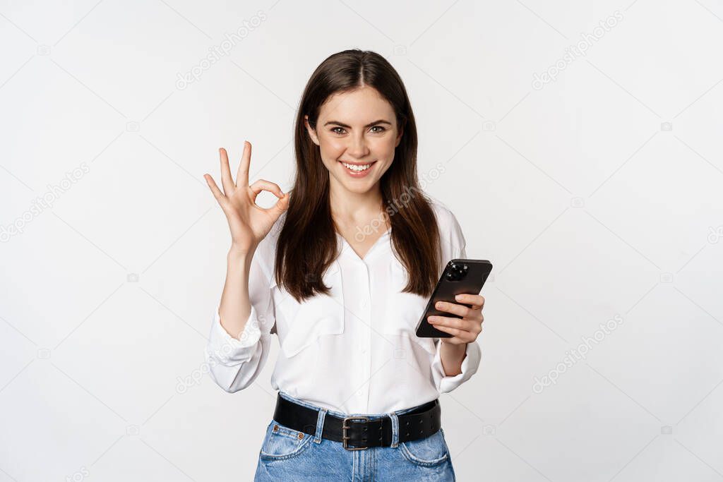 Smiling woman showing okay, ok sign while using mobile phone, approve app on smartphone, excellent service, online store, standing over white background