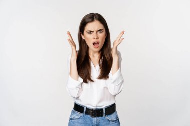 Frustrated and angry young woman gasping, looking shocked and upset at camera, offended by smth, standing bothered against white background