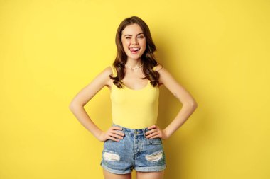 Stylish young brunette woman showing her white healthy teeth, smiling and showing tongue, winking enthusiastic, standing over yellow background