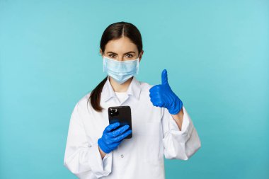 Online doctor and clinic. Young woman in medical face mask, using smartphone for client remote online appointment, showing thumbs up, standing over blue background
