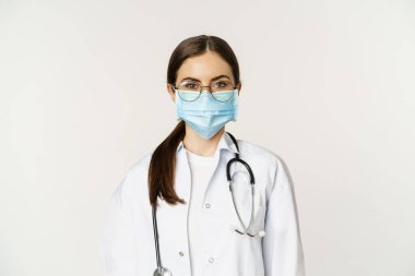 Close up portrait of female doctor, hospital staff in glasses and medical face mask, looking serious, ready to greet hospital patients, standing over white background