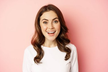 Close up of enthusiastic brunette girl with makeup, smiling and looking happy at camera, posing against pink background
