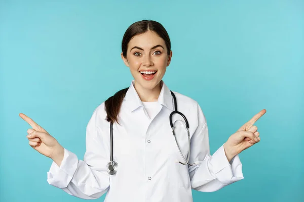Enthusiastic woman doctor, medical worker in white coat, pointing fingers sidweays, left and right, showing health clinic promo, standing over torquoise background — Stok fotoğraf