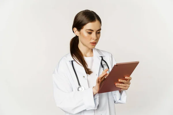 Woman doctor looking concerned at digital tablet, reading with worried face expression, wearing white coat, standing over white background — Foto de Stock