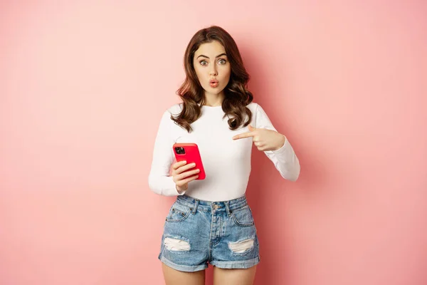 Cute girl pointing finger at smartphone with curious face expression, have you seen this gesture, showing online shopping offer on mobile phone app, pink background — Foto Stock