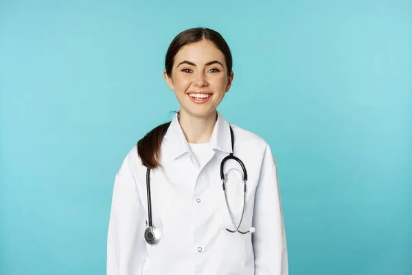 Smiling healthy medical worker, young woman doctor looking happy, standing in white coat and stethoscope against blue background — Zdjęcie stockowe