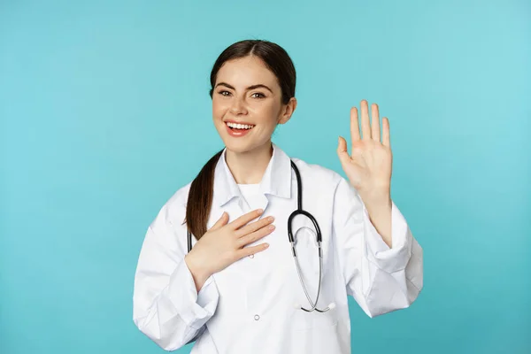 Friendly smiling woman doctor raising hand, name herself, introducing, standing in lab white coat against torquoise background — ストック写真