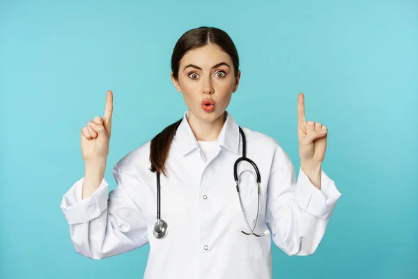 Portrait of surprised young doctor, female medical worker pointing fingers up, gasping amazed, showing health advertisement, standing over torquoise background — Stok fotoğraf
