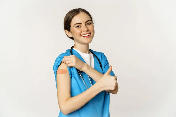 ワクチン接種後の彼女の肩を示す幸せな笑顔の医療従事者、医師または看護師、 covid-19ワクチンと親指を満足し、白い背景の上に立って — ストック写真